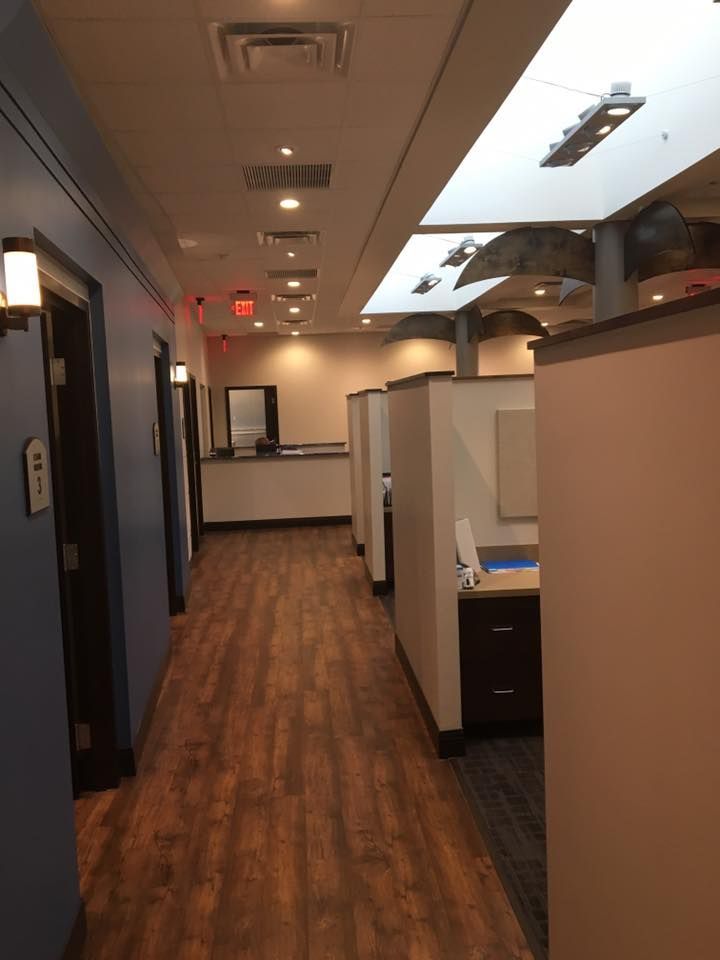 Hallway in a medical office, with exam rooms on one side and workstations on the other, wood flooring, and bright lighting.