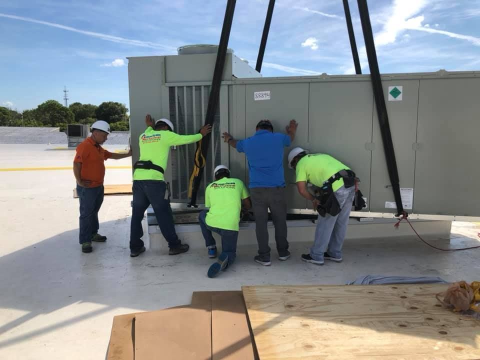 Five workers lifting an HVAC unit on a rooftop. Workers in neon green and blue shirts guide the unit with straps.