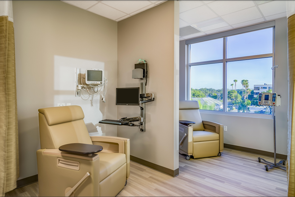 Chemotherapy room with recliner, medical equipment, and large window overlooking trees.
