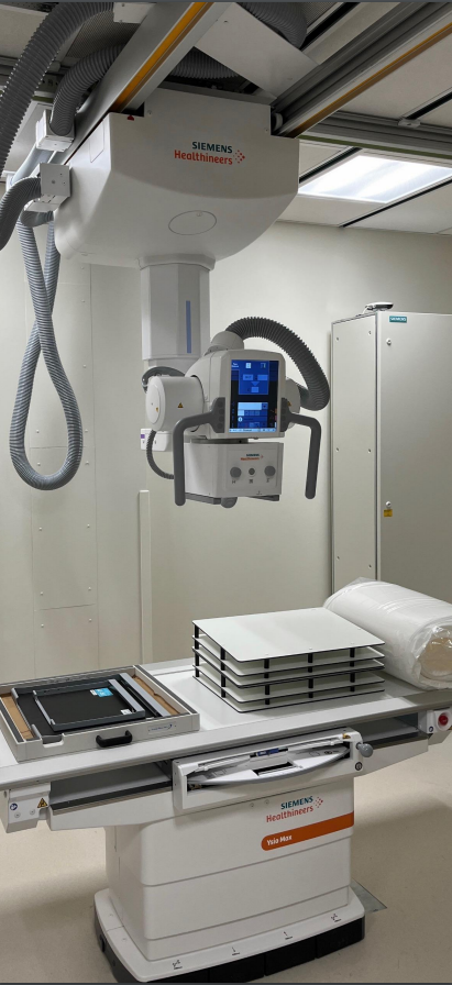 An X-ray machine in a medical room. The machine is white with orange accents.