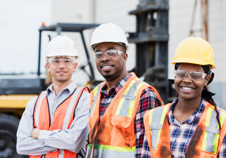 Three construction workers wearing hard hats, safety glasses, and vests smile for the camera.
