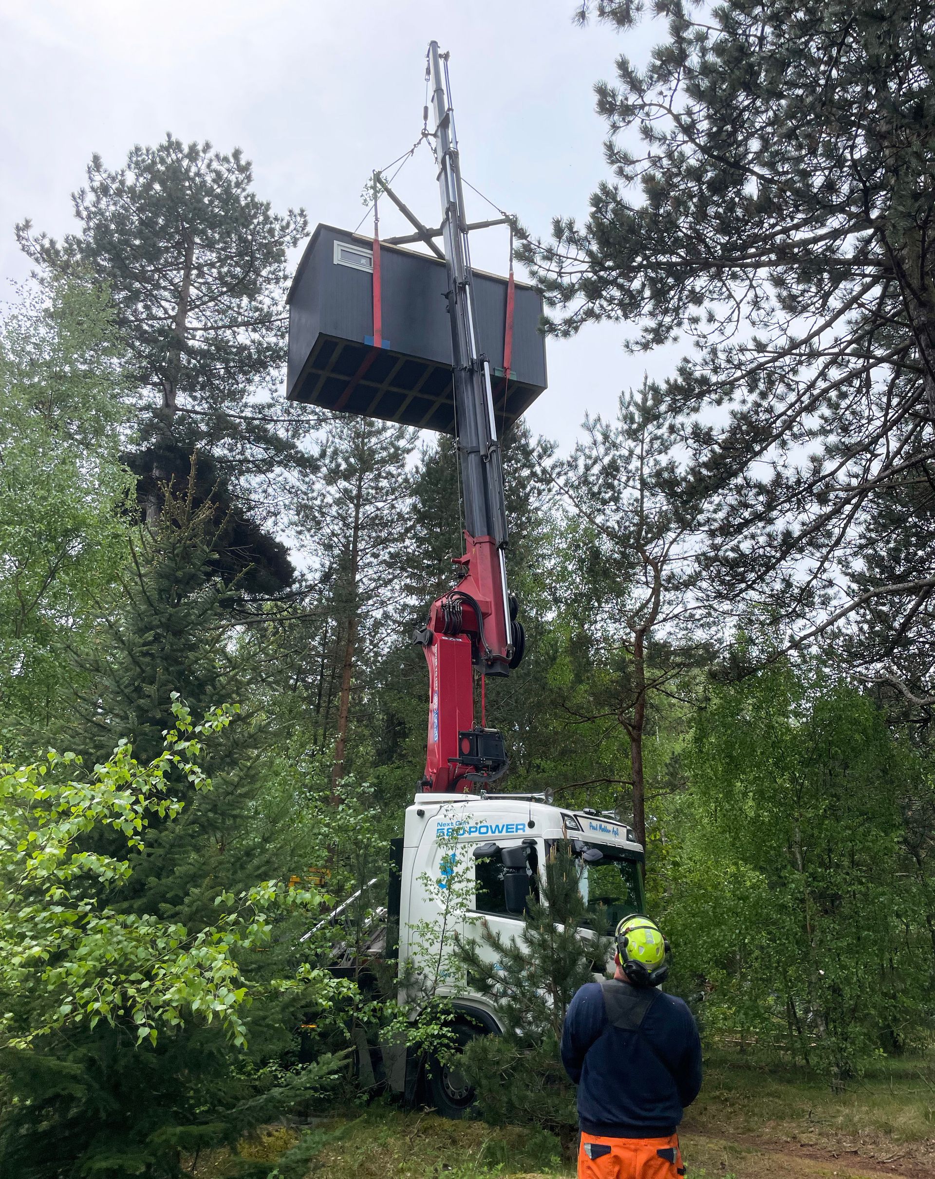 En mand står ved siden af ​​en lastbil med en kran knyttet til den, som løfter en skurvogn.
