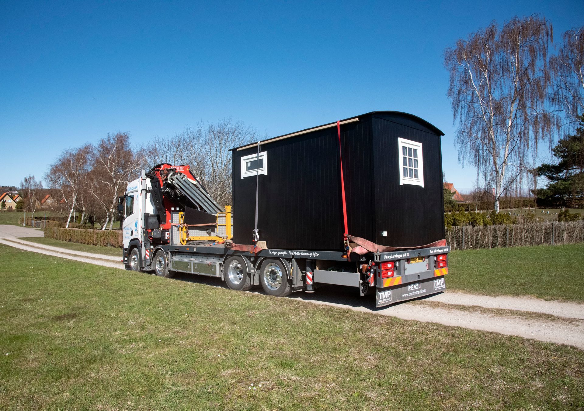 En lastbil transporterer et lille sort hus på en grusvej.