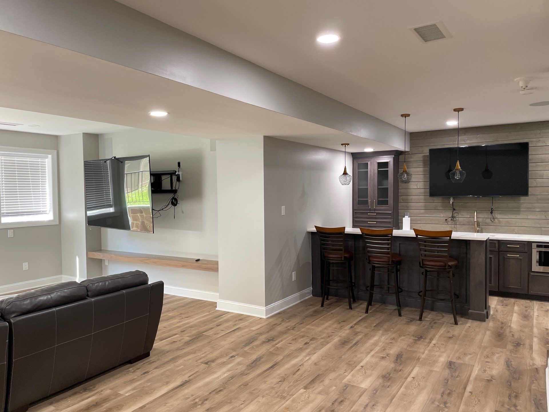Finished basement with a bar, TV, and seating. Wooden floors and gray walls.