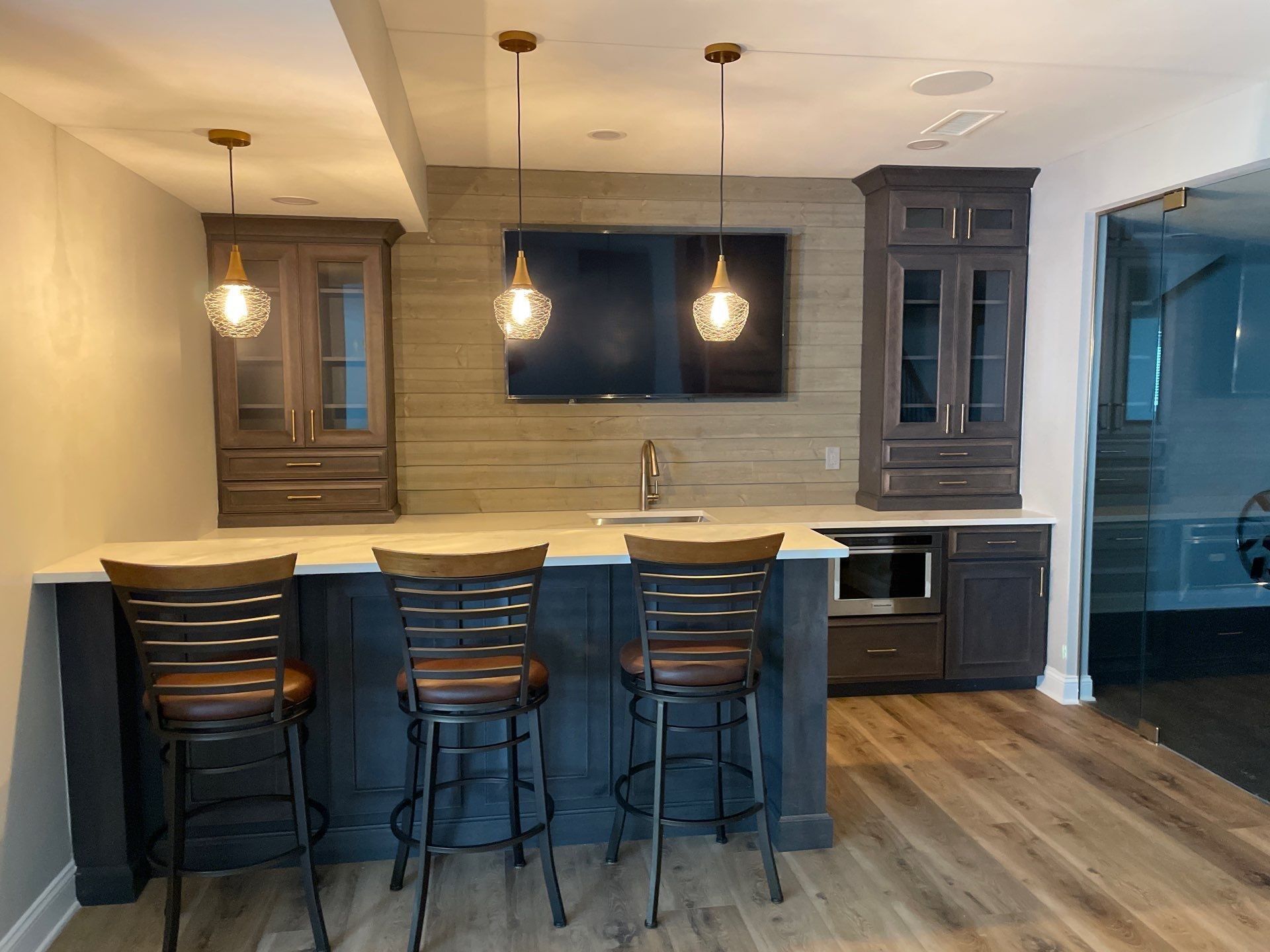 Basement bar with dark cabinets, pendant lights, seating, and a TV on a textured wall.