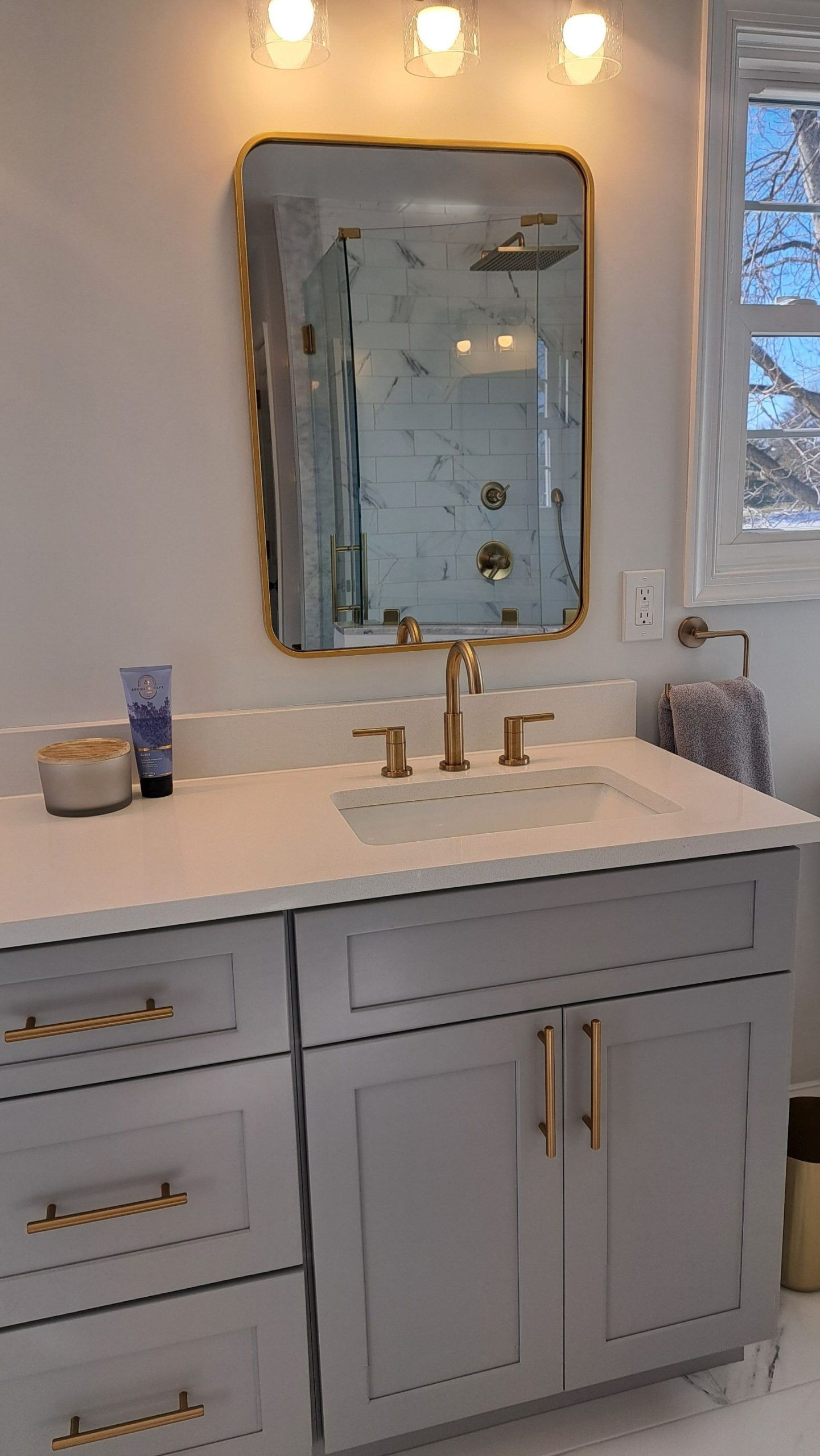 Bathroom vanity with gold-framed mirror, gray cabinet, white countertop, gold fixtures, and a window.
