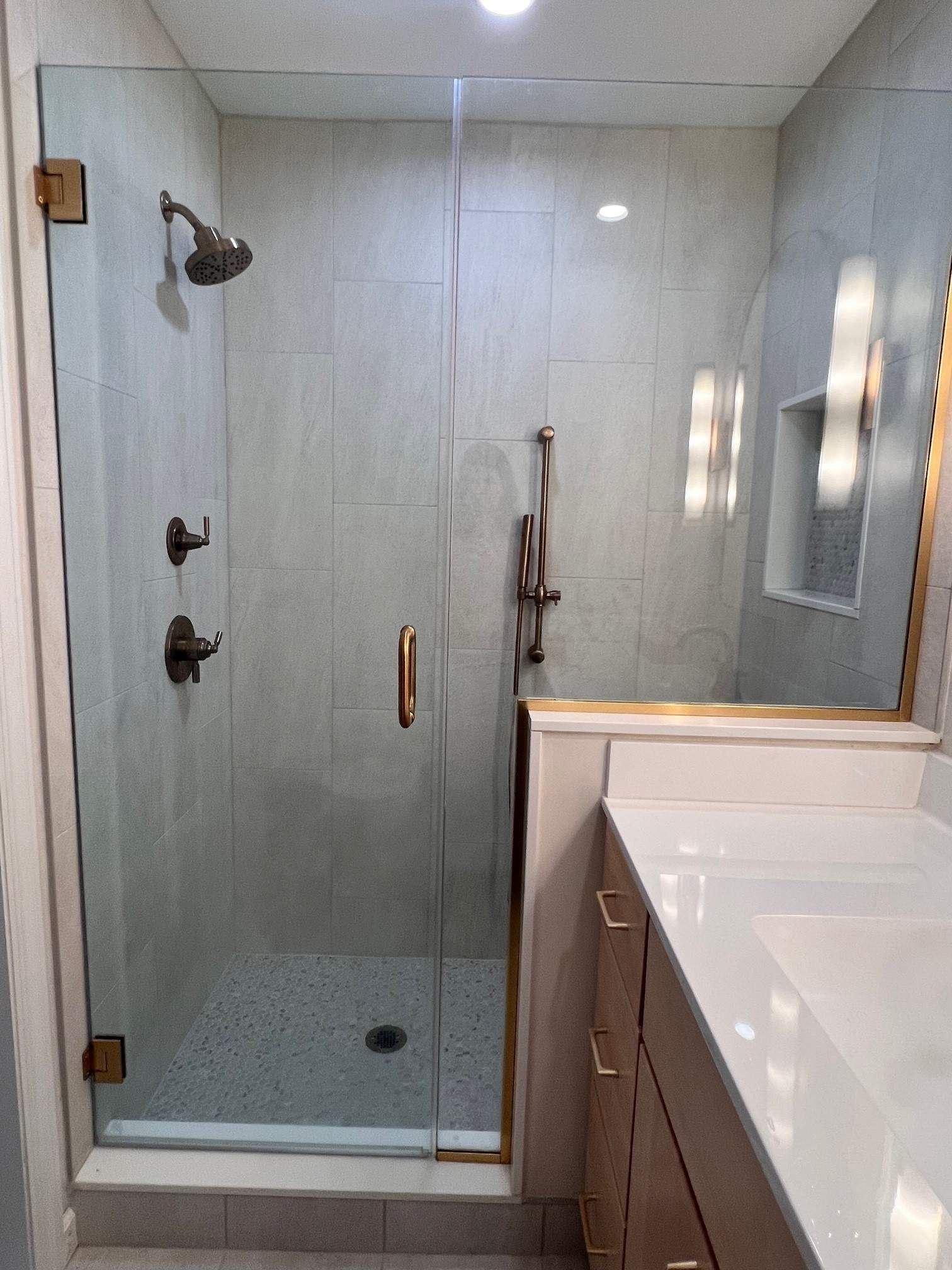 A glass shower with bronze fixtures and light gray tiled walls, next to a white vanity.
