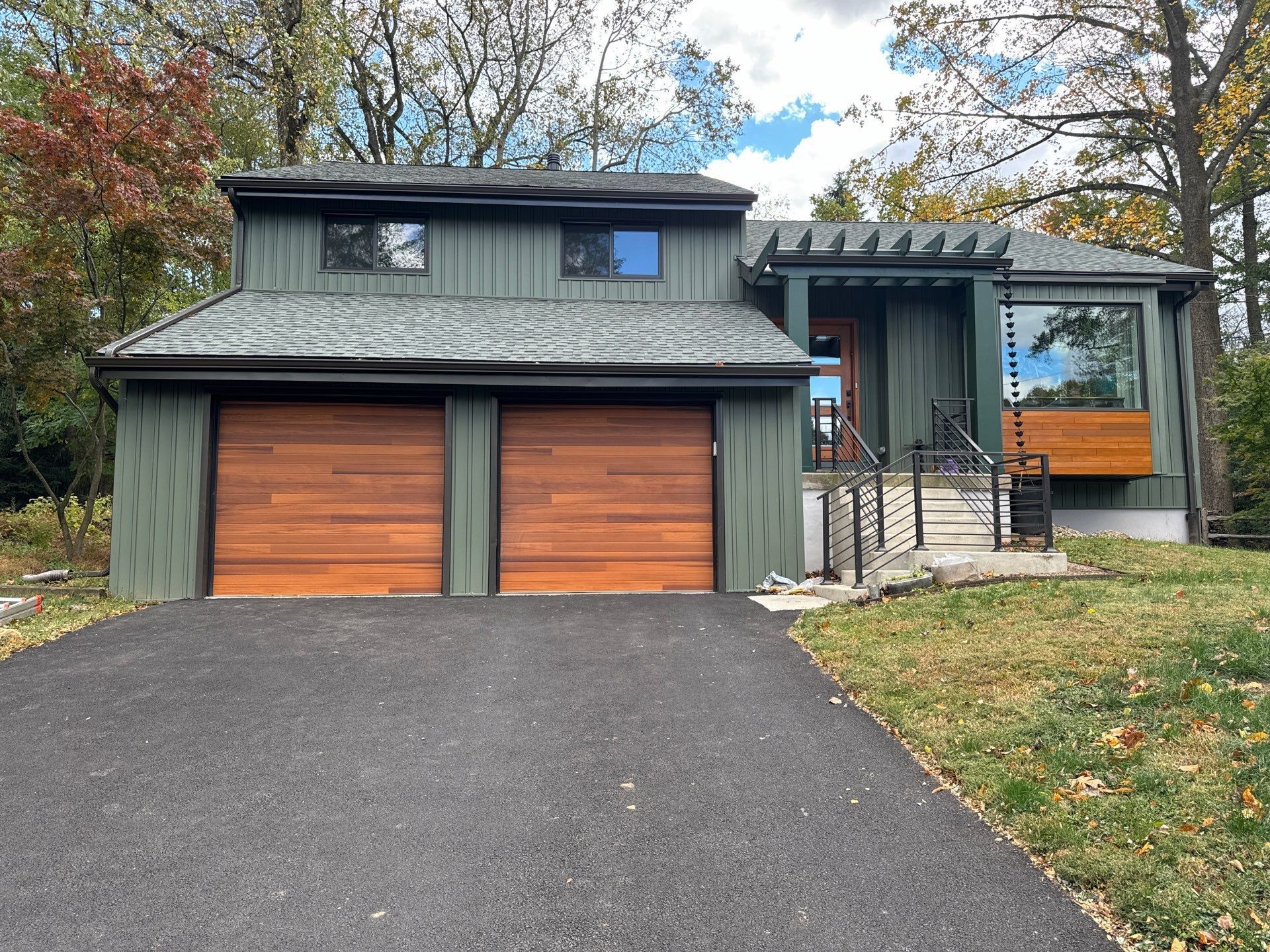 A green house with two wooden garage doors, a black driveway, and a window with a wooden frame.