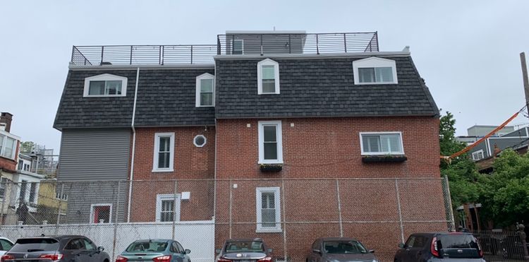 Red brick building with a dark roof and a rooftop deck. Cars are parked in front of the building.