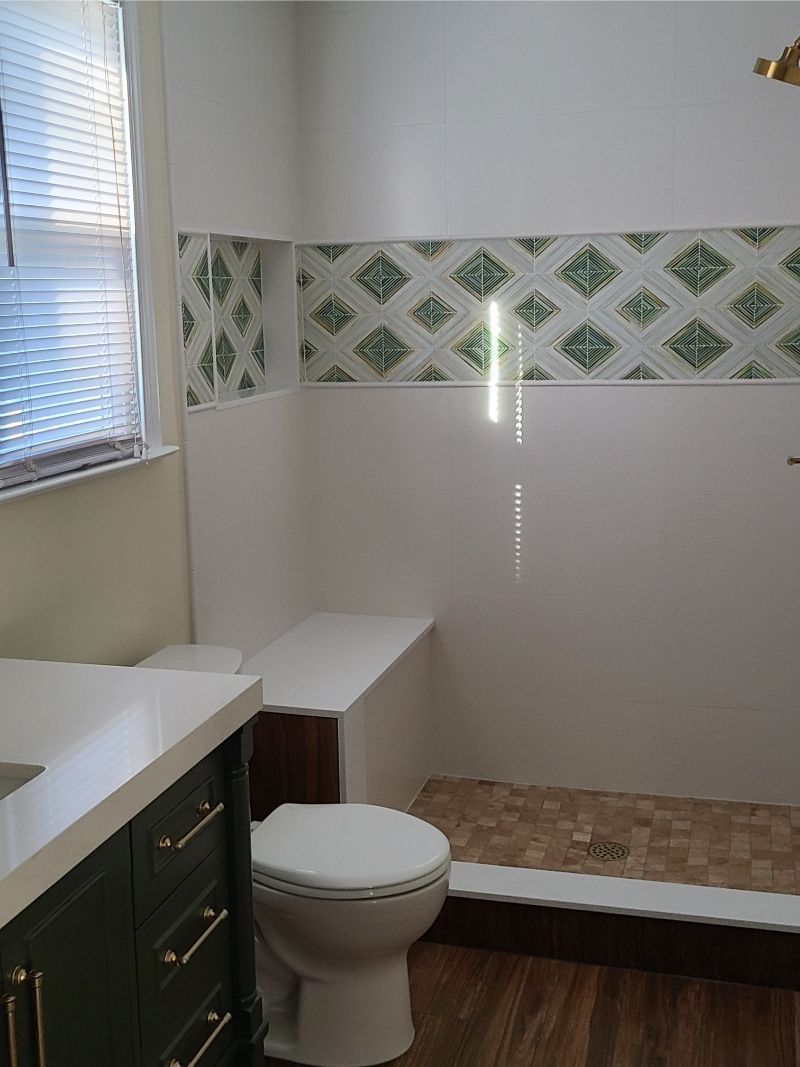 Bathroom with white walls, patterned green tile, toilet, vanity, and shower with a bench.
