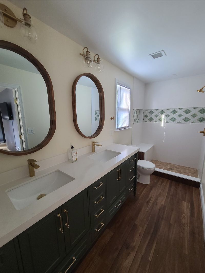 Bathroom with double sink vanity, oval mirrors, gold fixtures, and wood-look flooring.
