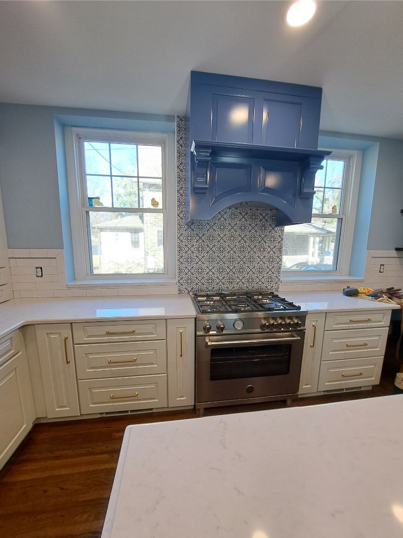 Kitchen with blue range hood, stove, mosaic backsplash, and white cabinets.
