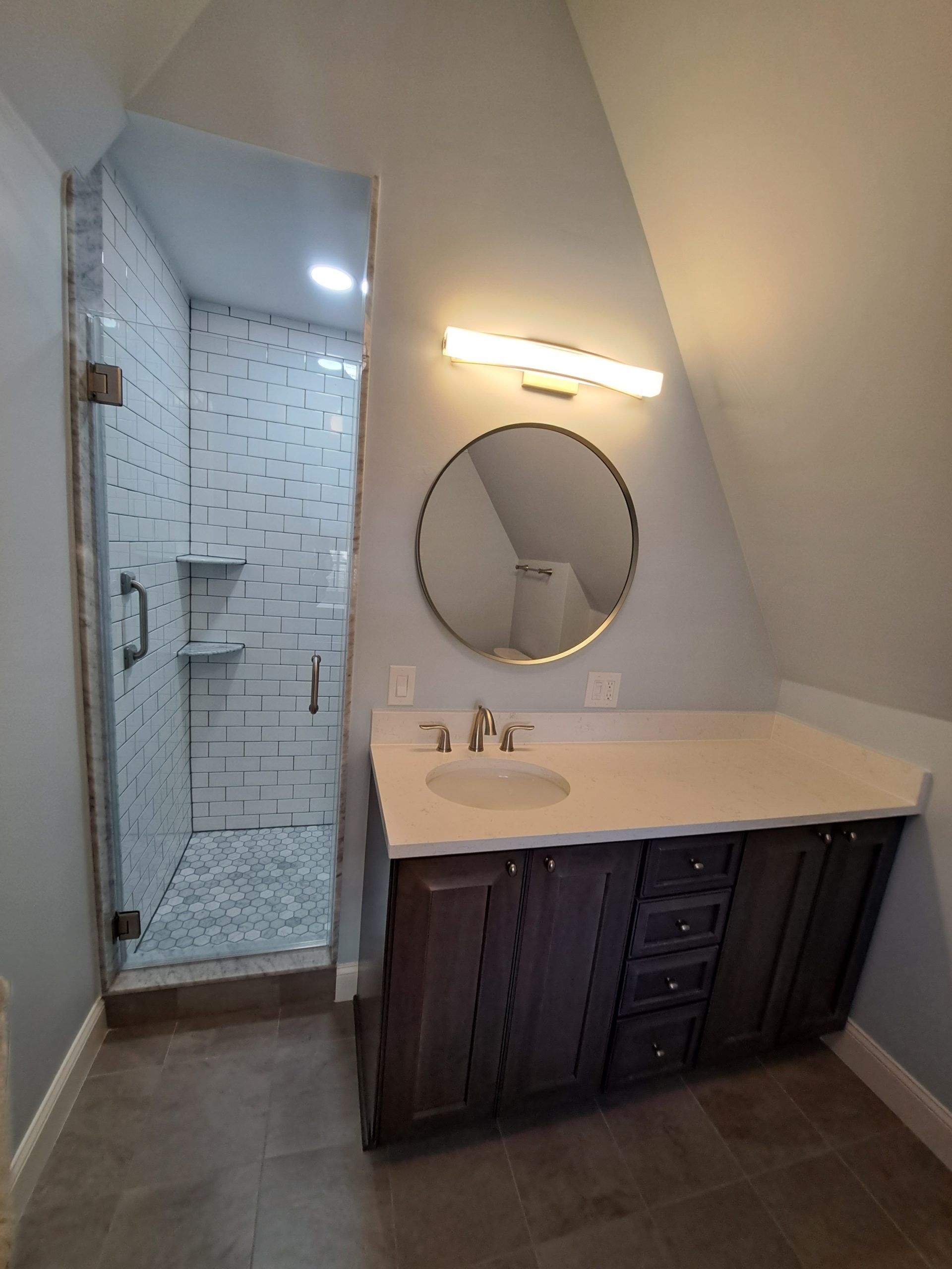 Bathroom with shower and vanity. Brown cabinets, white countertop, round mirror, angled ceiling.