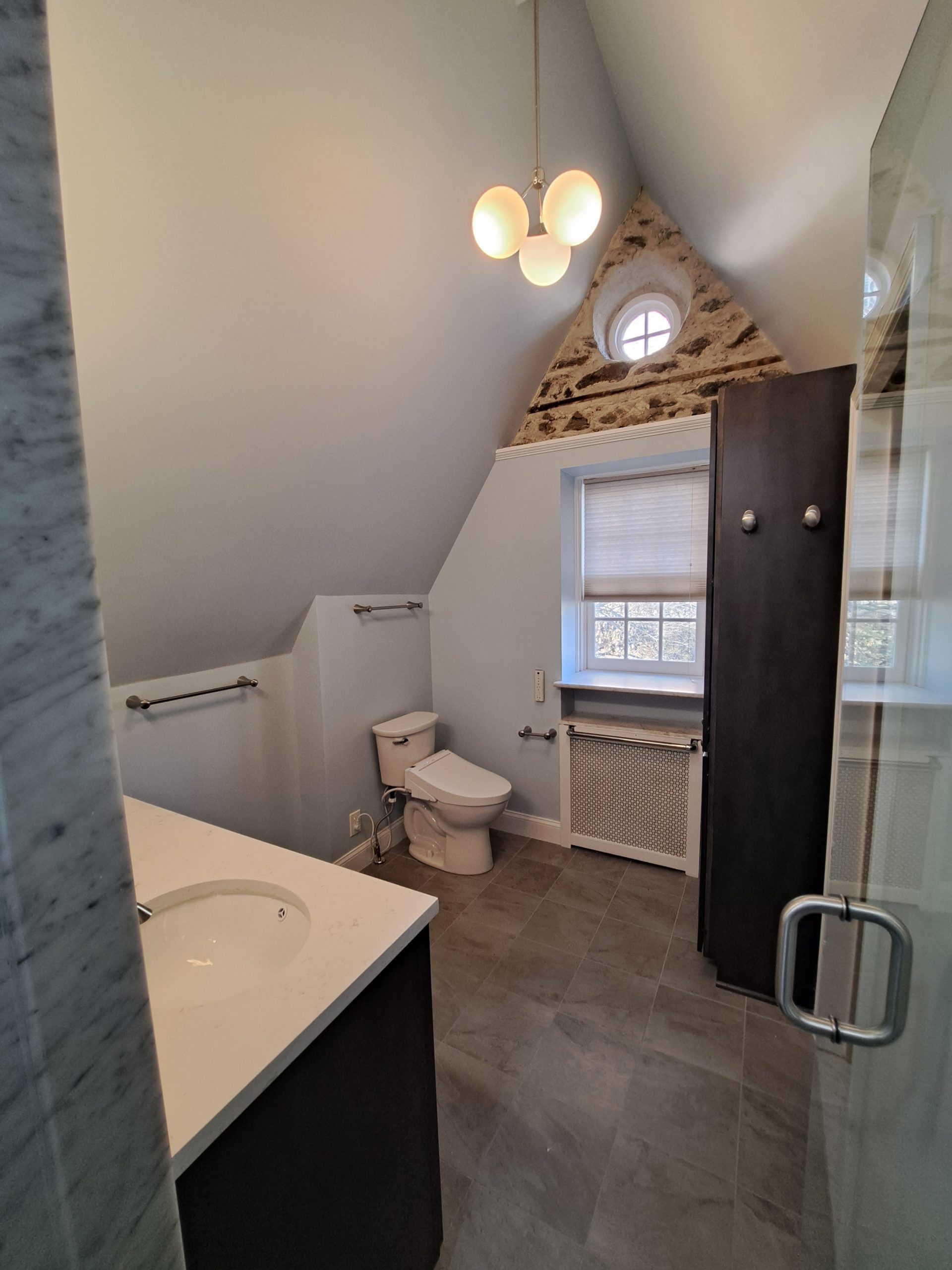 Bathroom with sloped ceiling, vanity, toilet, window, and dark wood cabinet. Stone accent wall.