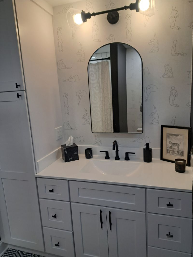 Bathroom vanity with white cabinets, black hardware, arched mirror, black faucet, and patterned wallpaper.