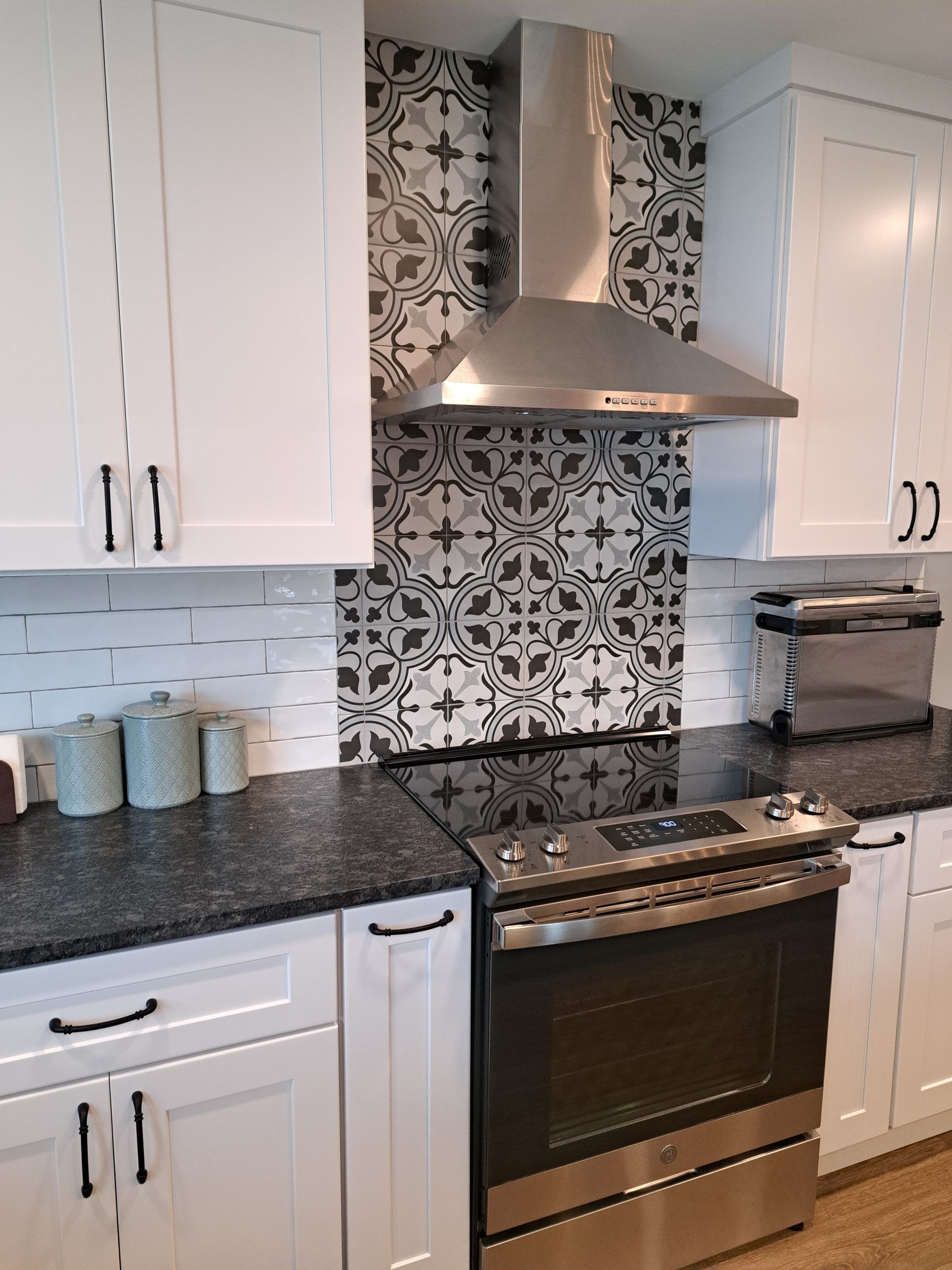 White kitchen with a patterned backsplash, stainless steel range hood, and black countertops.