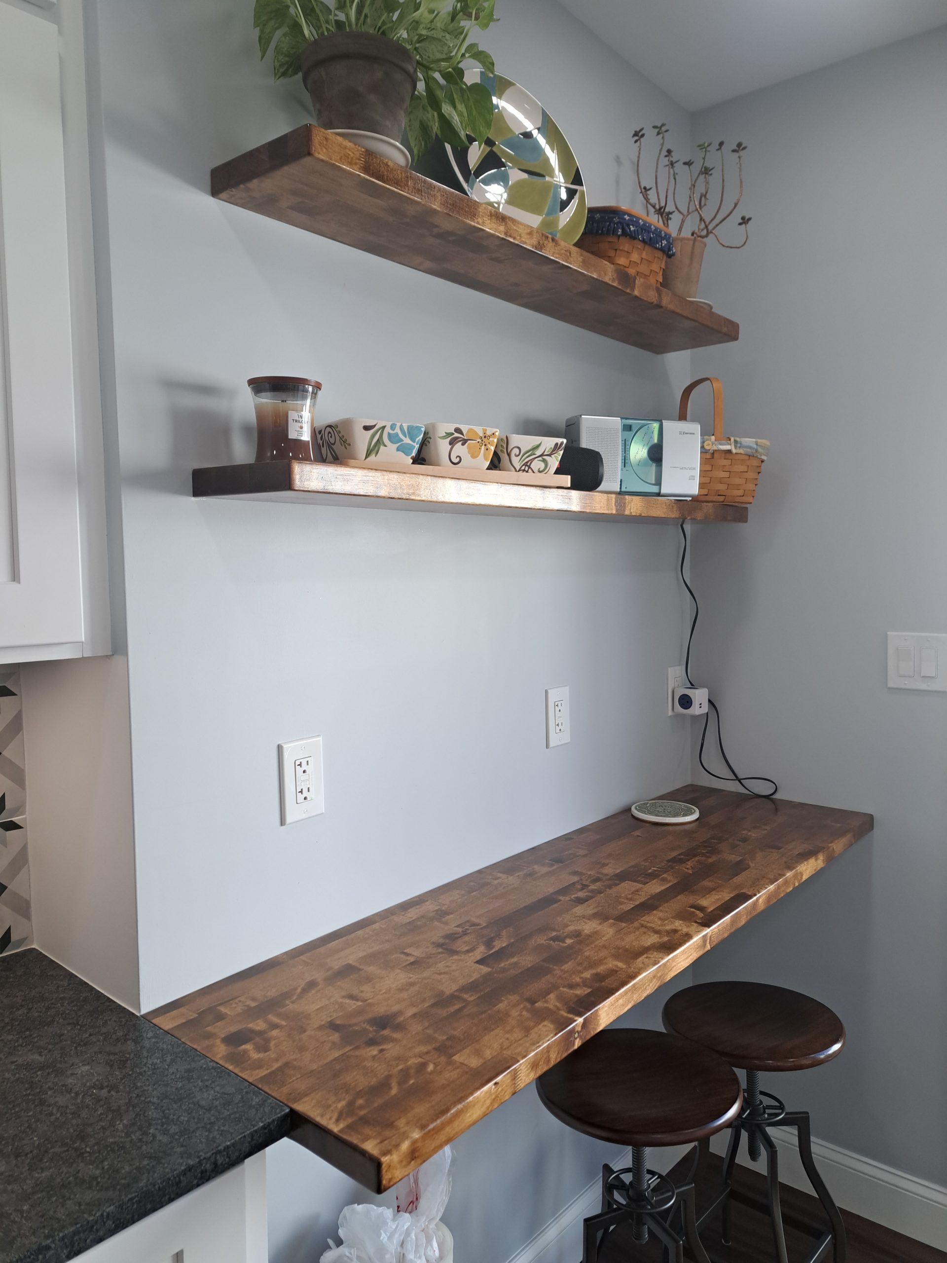 Wooden shelves and countertop in a kitchen. Items on shelves, two stools. Blue-gray wall.