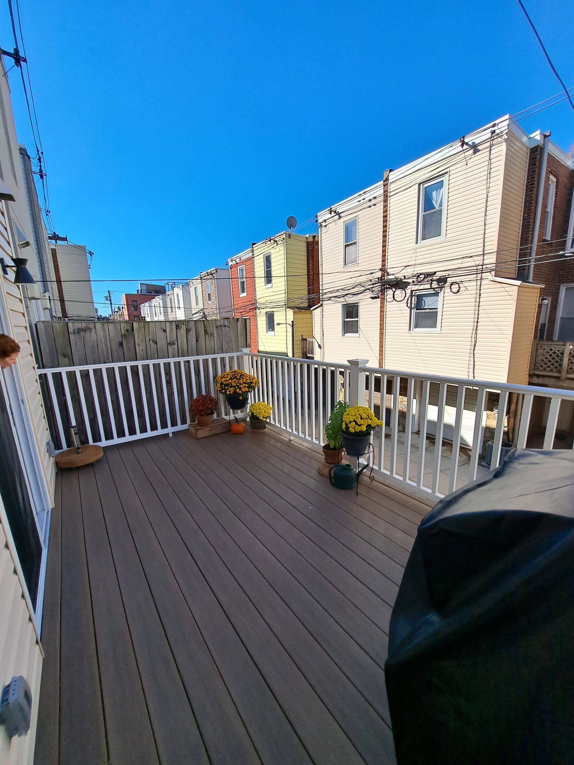 Wooden deck with white railing, potted flowers, and view of urban buildings under a blue sky.