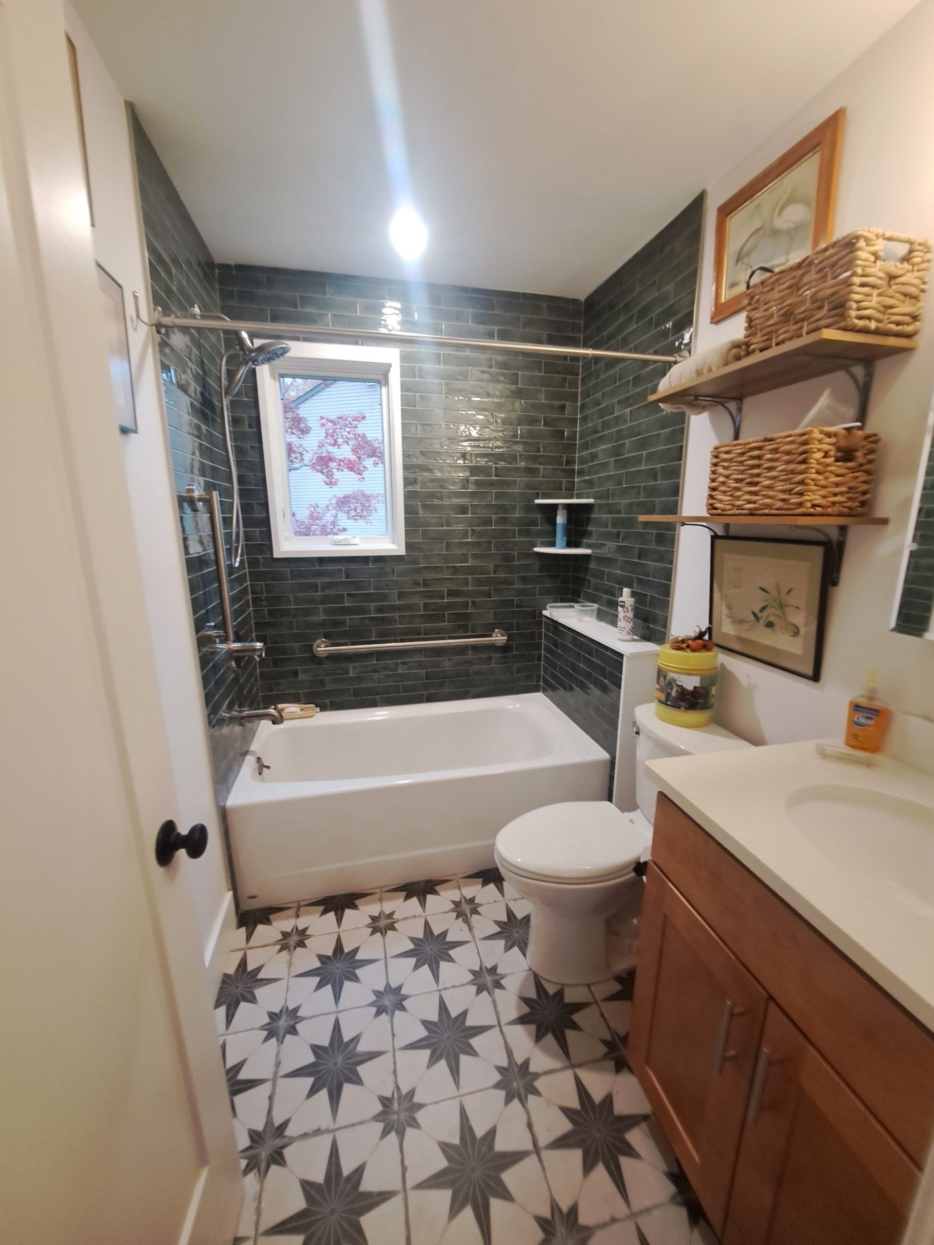 Small bathroom with dark green tiled shower, white tub, patterned floor, and wooden vanity.
