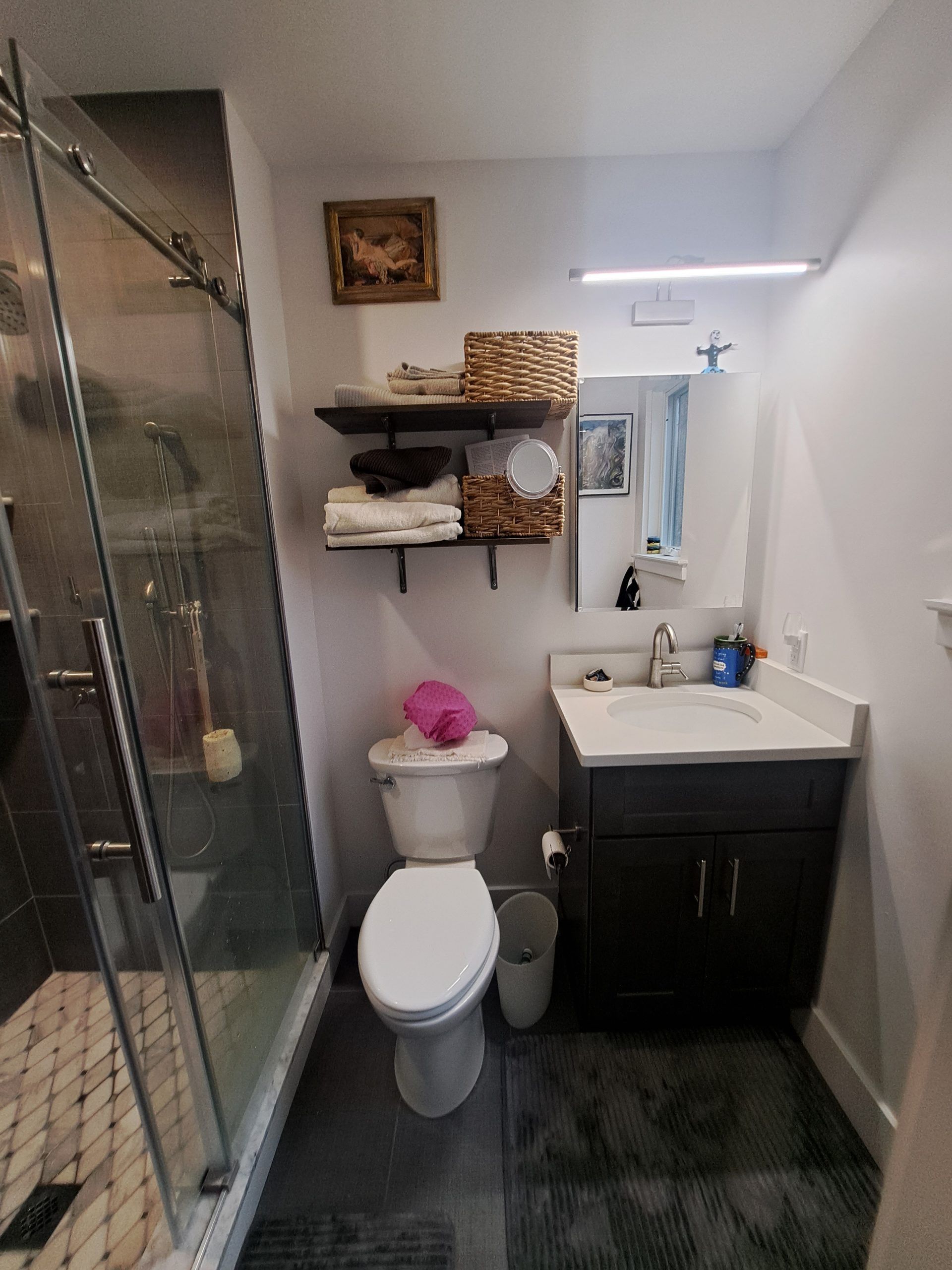 Bathroom with glass shower, toilet, dark vanity, and shelving. Gray walls, dark flooring, and baskets.