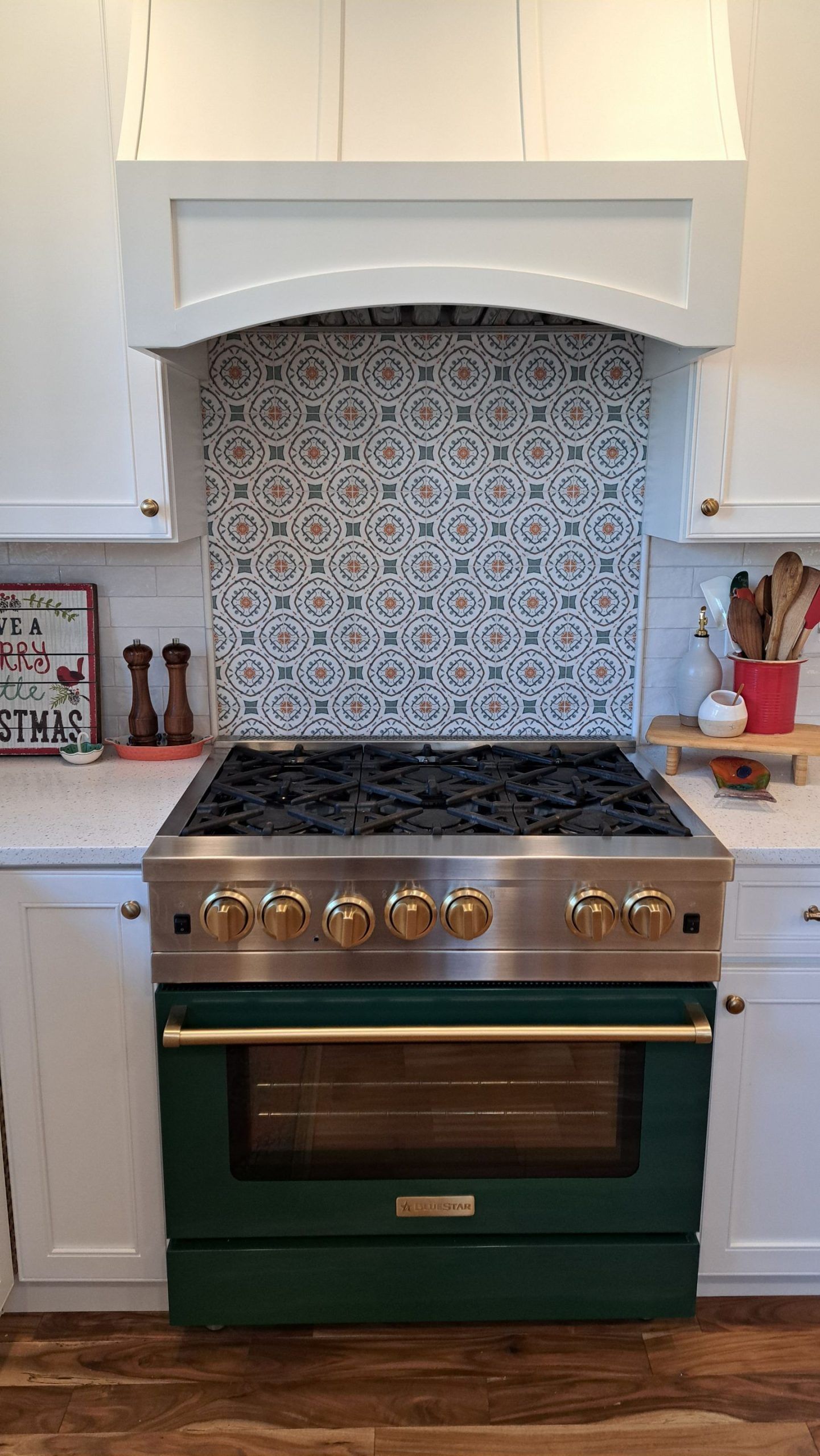 Green and gold stove with patterned backsplash, white range hood, and cabinets.