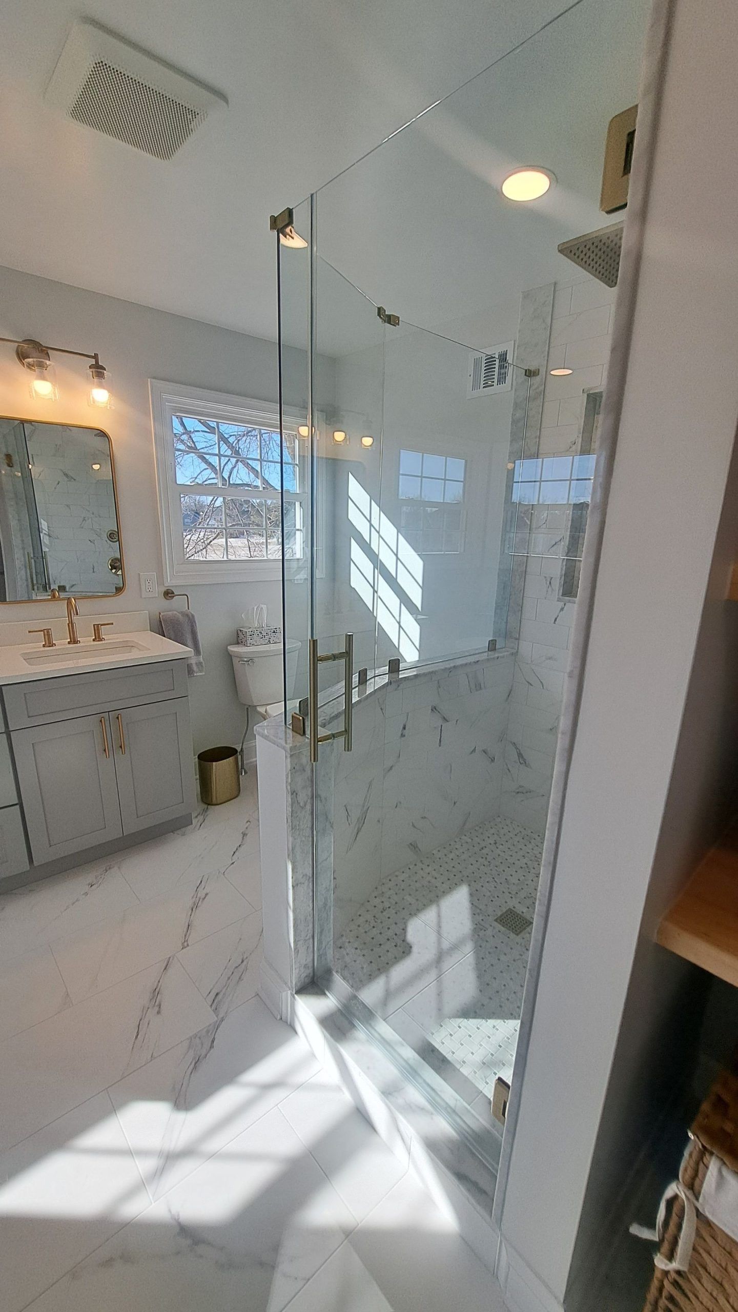 Bright bathroom with glass shower, white marble floors and walls, vanity, and window.