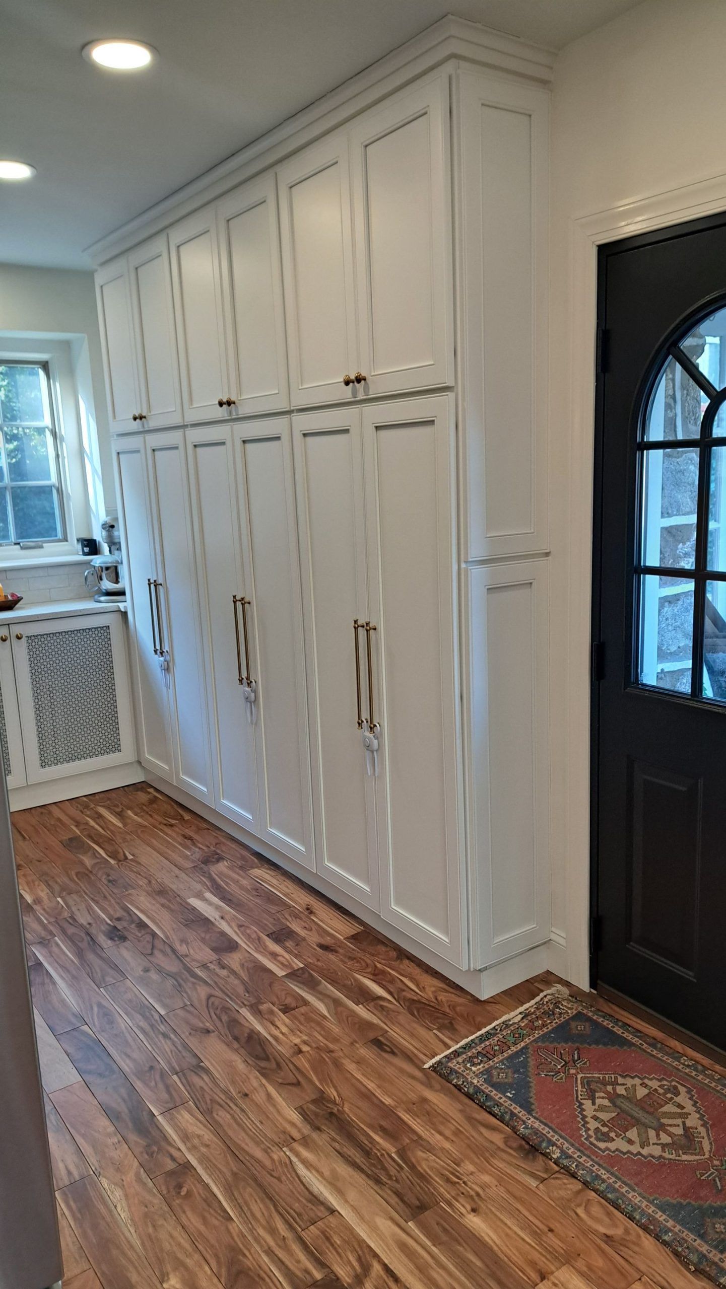 Tall white cabinets with brass handles line a hallway with wood flooring, next to a black door and window.