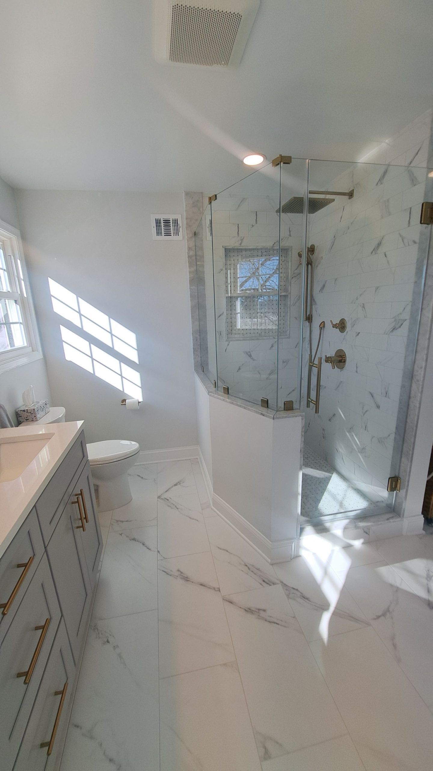 Bright, modern bathroom with marble tile, glass shower, vanity, and a window providing natural light.
