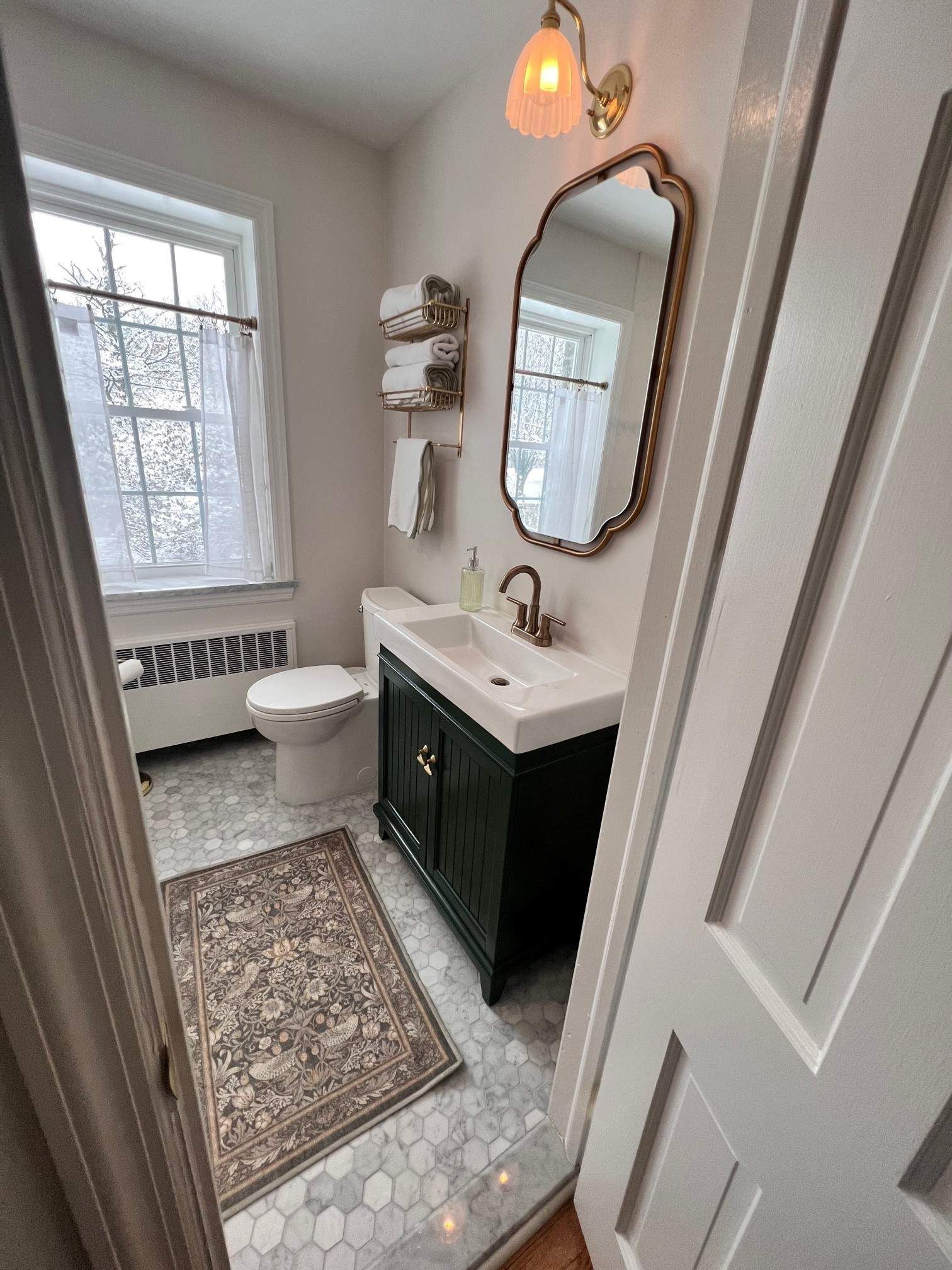 Small bathroom with dark green vanity, ornate gold mirror, window with sheer curtains, patterned rug.
