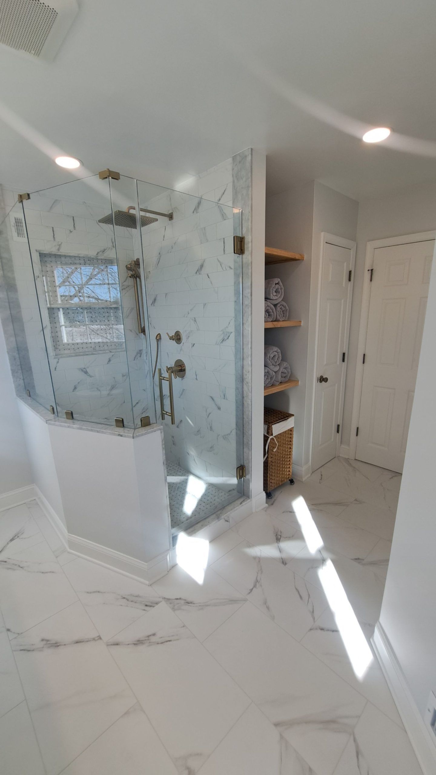 Modern bathroom with white marble tile, glass shower, and wooden shelves.