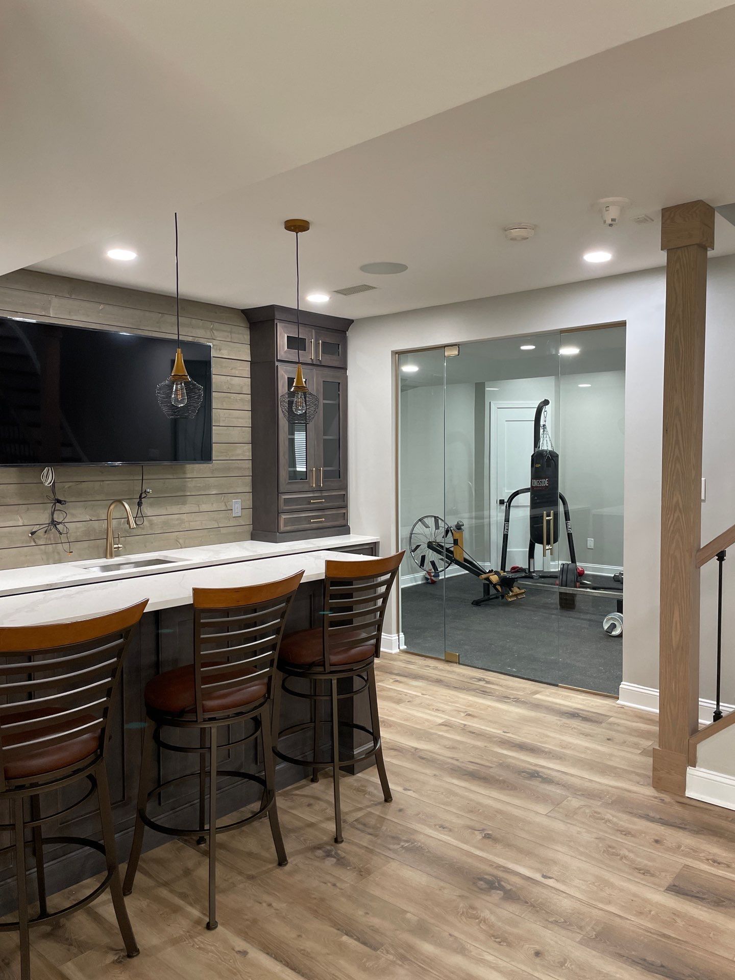 Basement with a bar, gym equipment visible through a doorway, and bar stools. Light wood flooring.