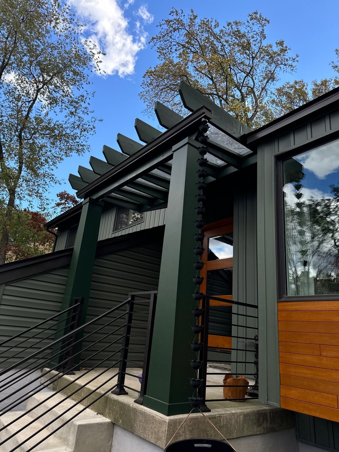 Green pergola and entrance with a dark door, steps, and black railing. Blue sky in the background.