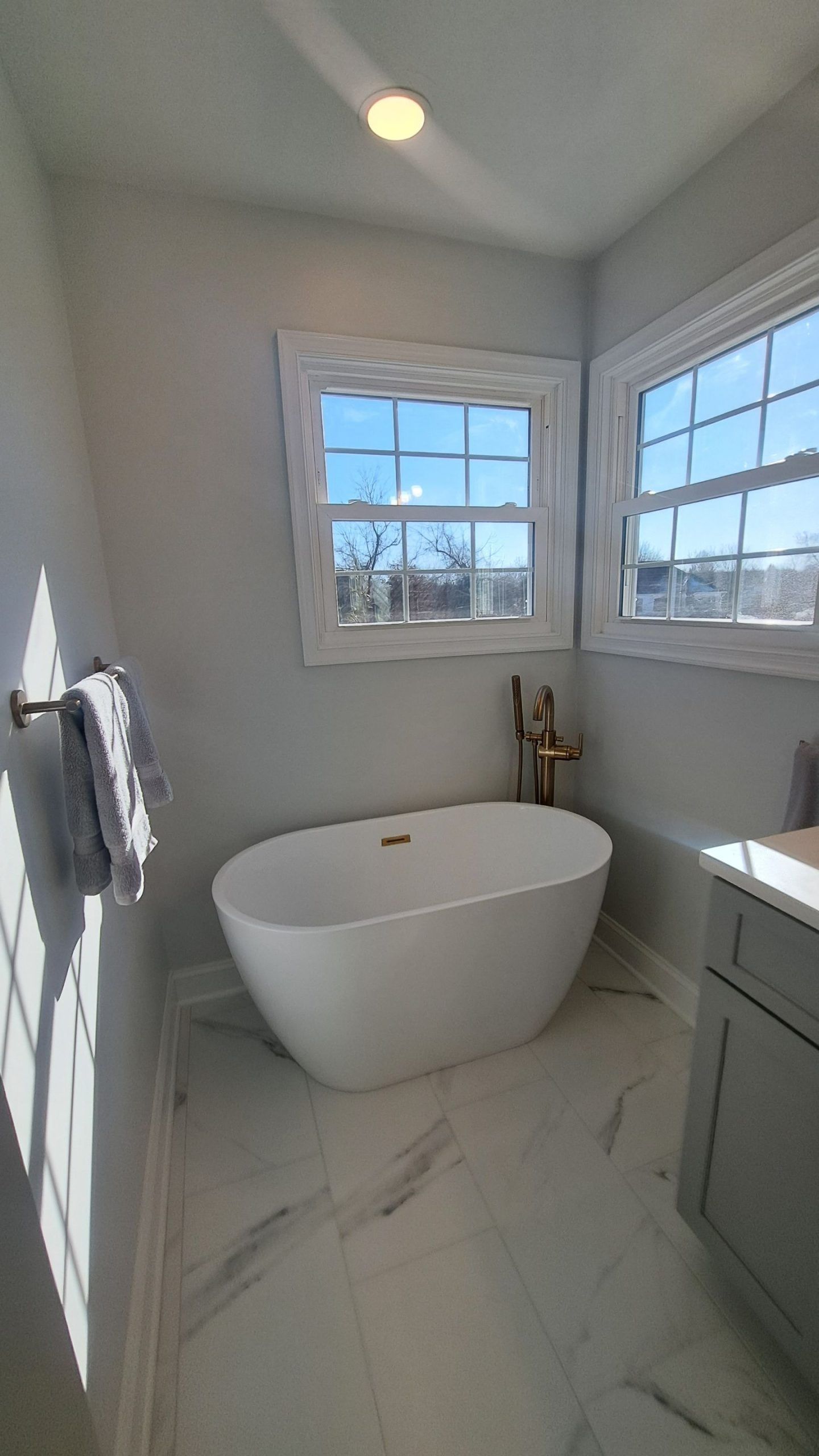 Modern bathroom with a white freestanding bathtub, two windows, and marble flooring.