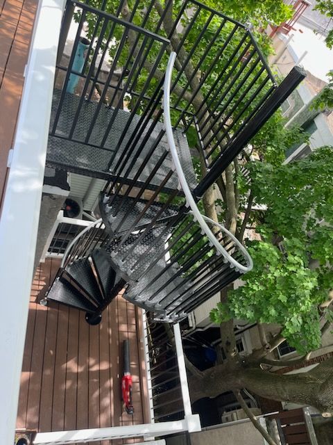 Spiral staircase with black metal railing outside a building with wooden decking and trees.