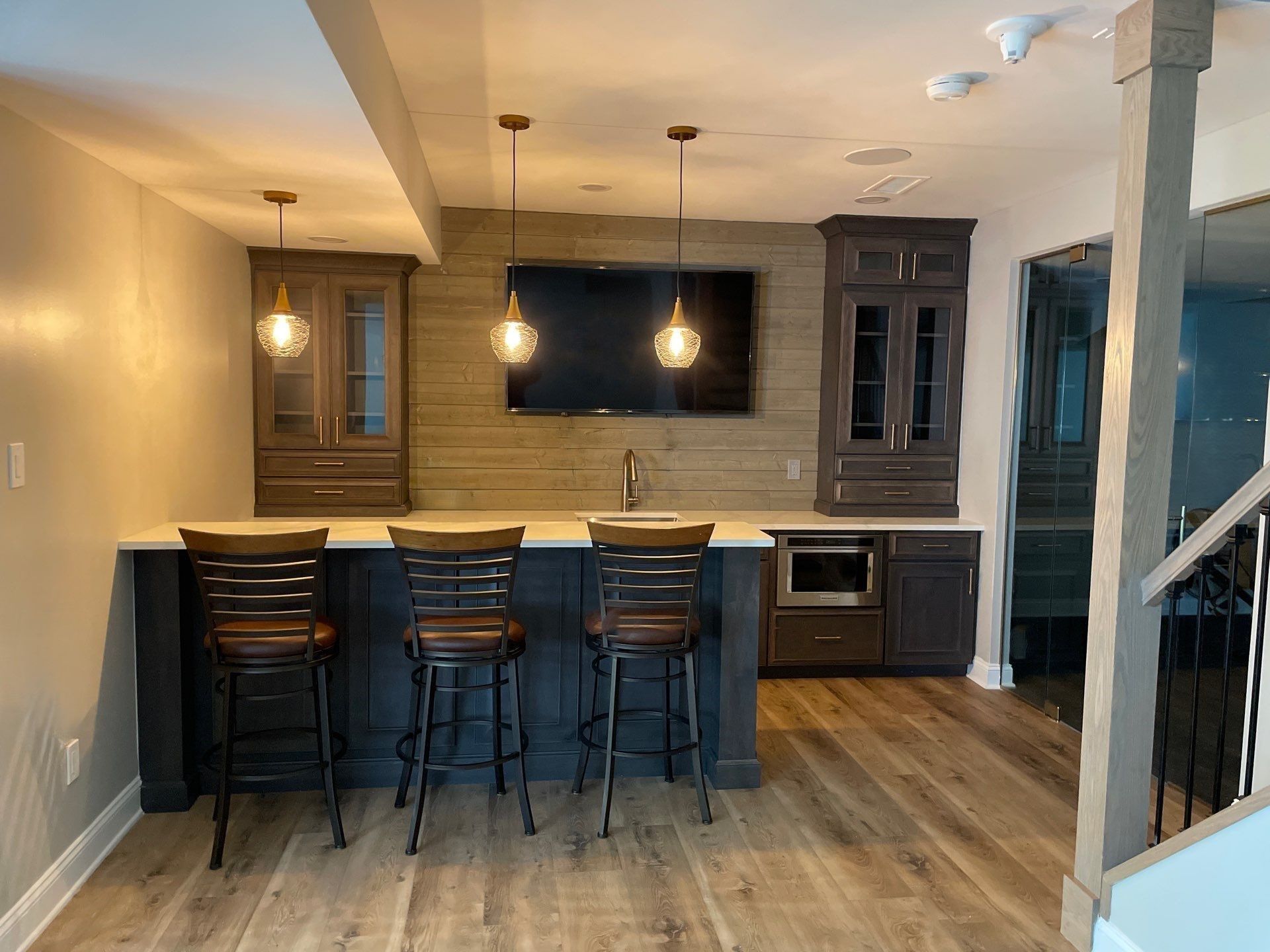 Basement bar with dark cabinets, countertop seating, TV, and pendant lights. Light wood flooring.