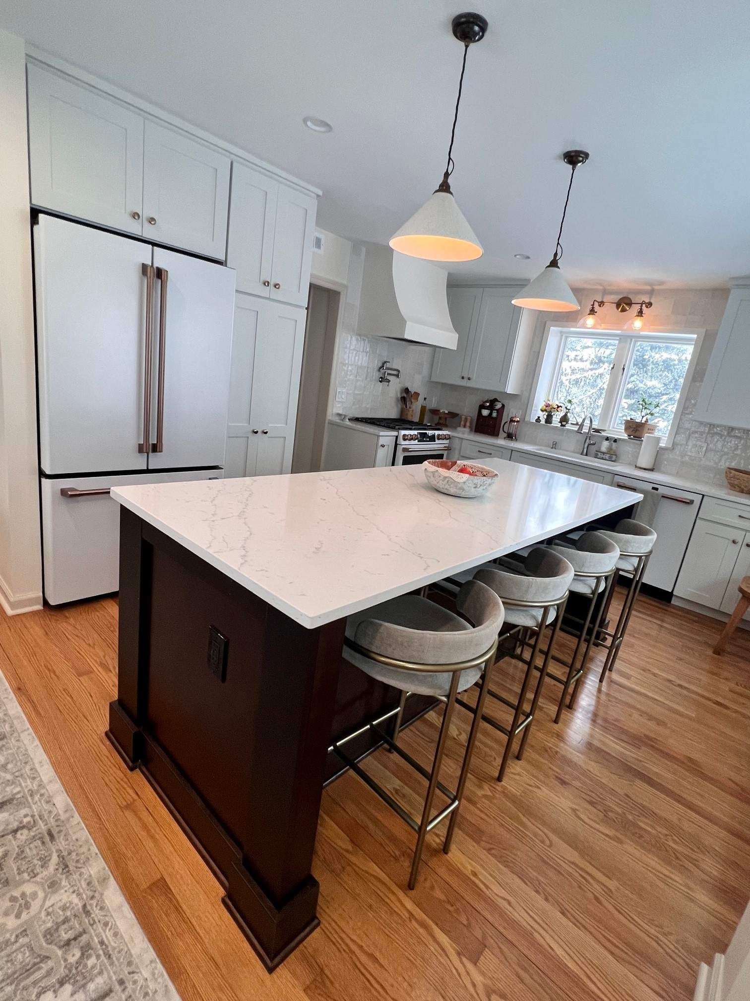 Modern kitchen with a dark wood island, white countertop, and bar stools. White cabinets and a refrigerator are visible.