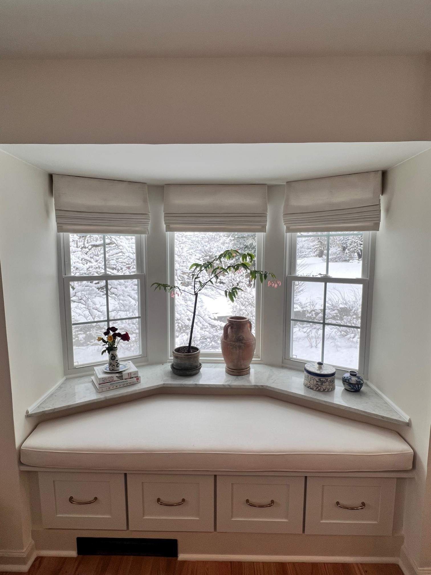 Bay window with a built-in bench, drawers, and neutral-colored shades. Snowy outside.