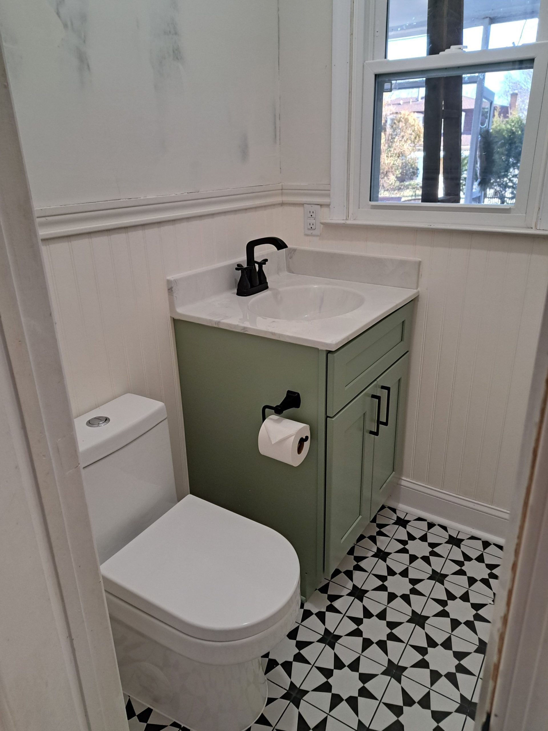 Small bathroom with green vanity, white toilet, patterned floor, and window.