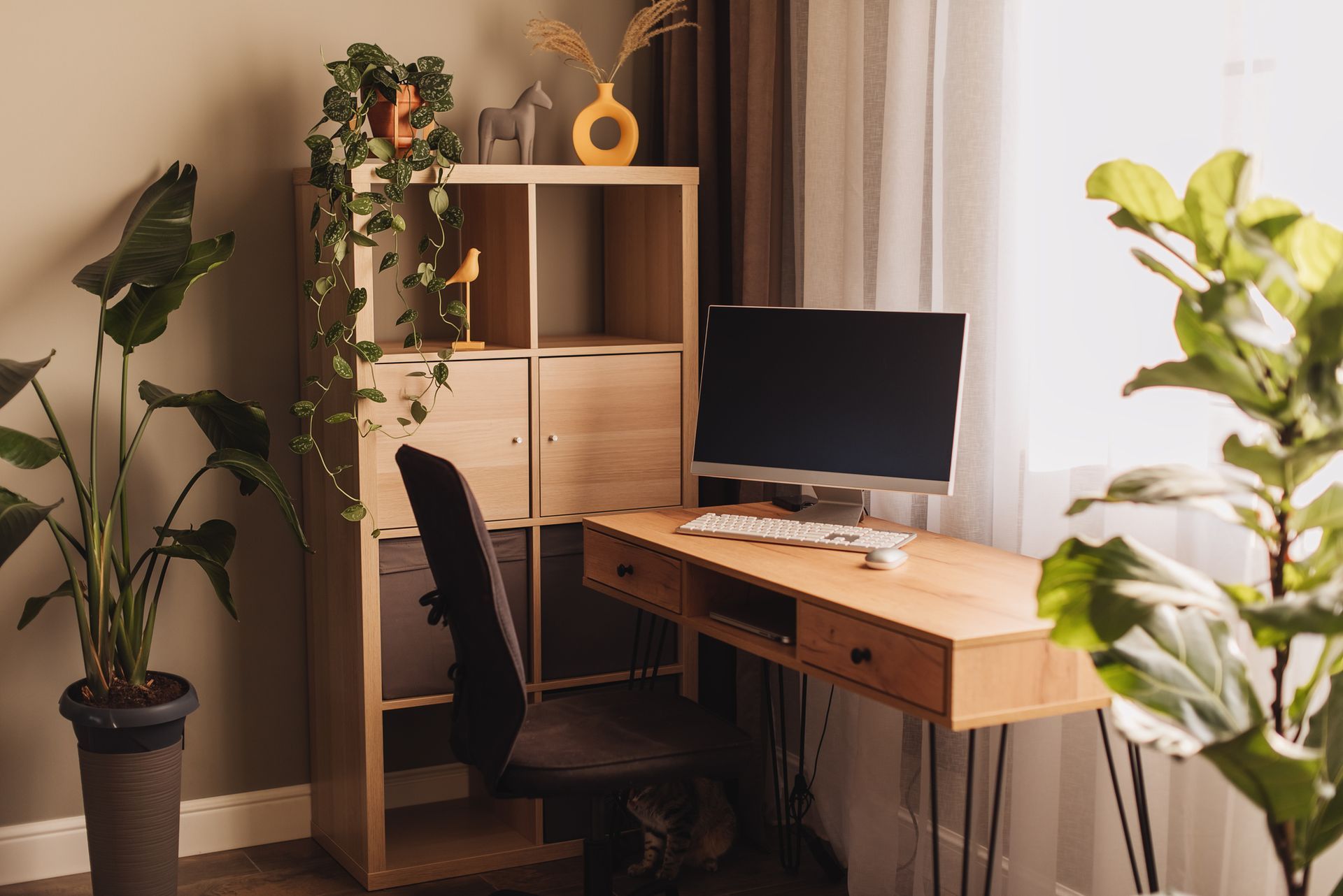Desk with computer, plants, and shelving unit by a window. Neutral tones.