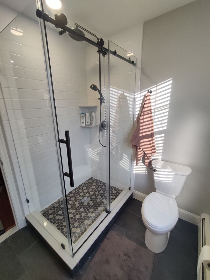 Shower with glass door, black hardware, white tile, pebble floor, towel on the wall, and toilet next to it.