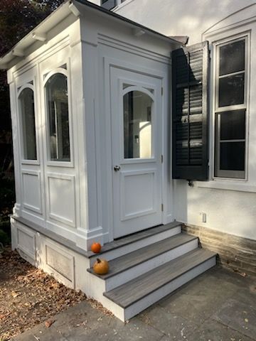 White, enclosed porch with steps, pumpkins, and a door, attached to a house with shuttered window.