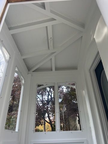 White porch ceiling with exposed beams, glass windows, and a doorway, trees reflected in the glass.