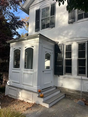White house with a small, enclosed porch, steps, and pumpkins. Black shutters and a bright, sunny day.
