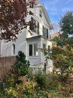 White two-story house with bay windows and dark shutters, surrounded by autumn trees and foliage.