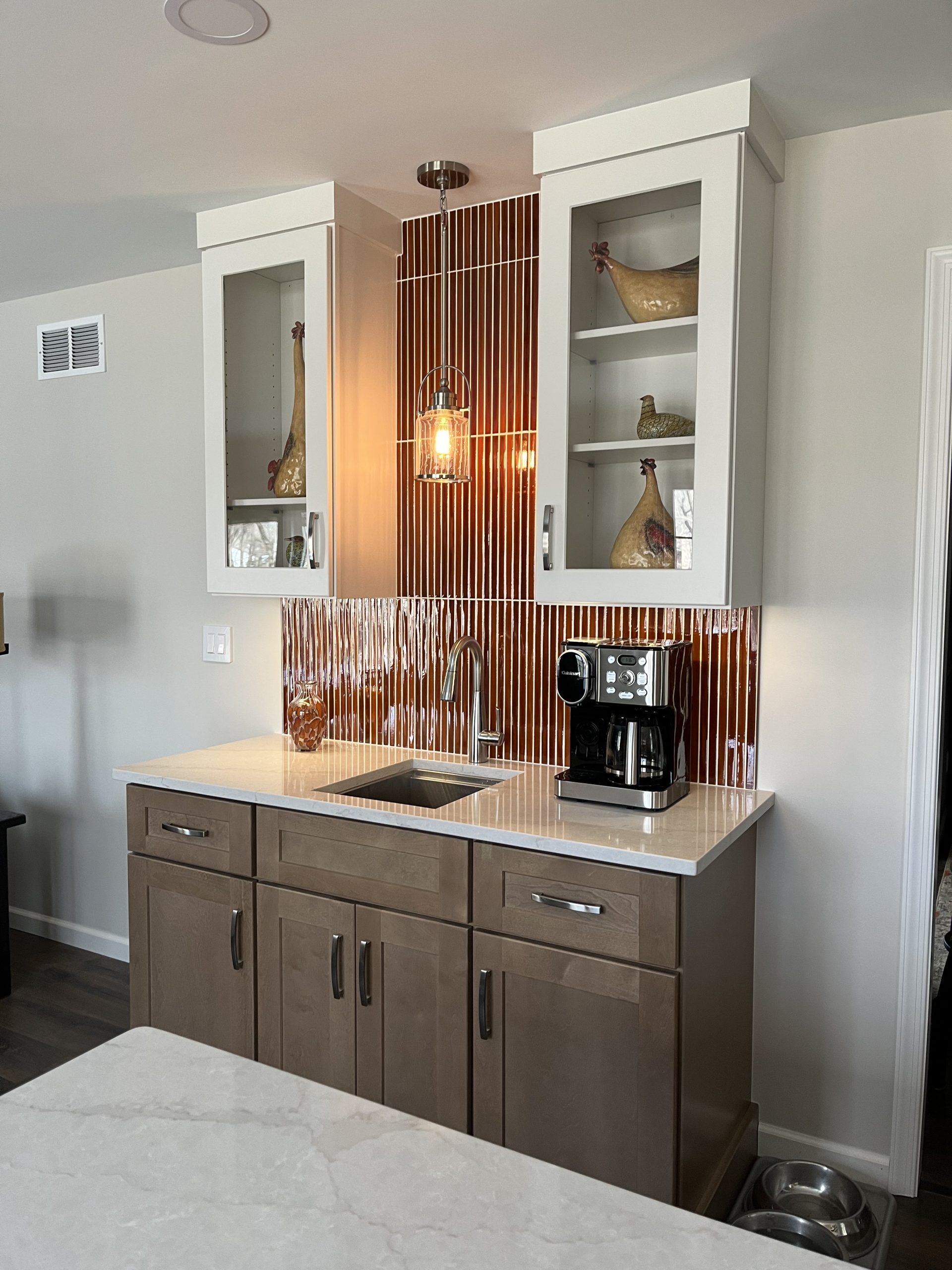 A coffee bar with upper glass-door cabinets, copper backsplash, and beige countertops, gray lower cabinets.