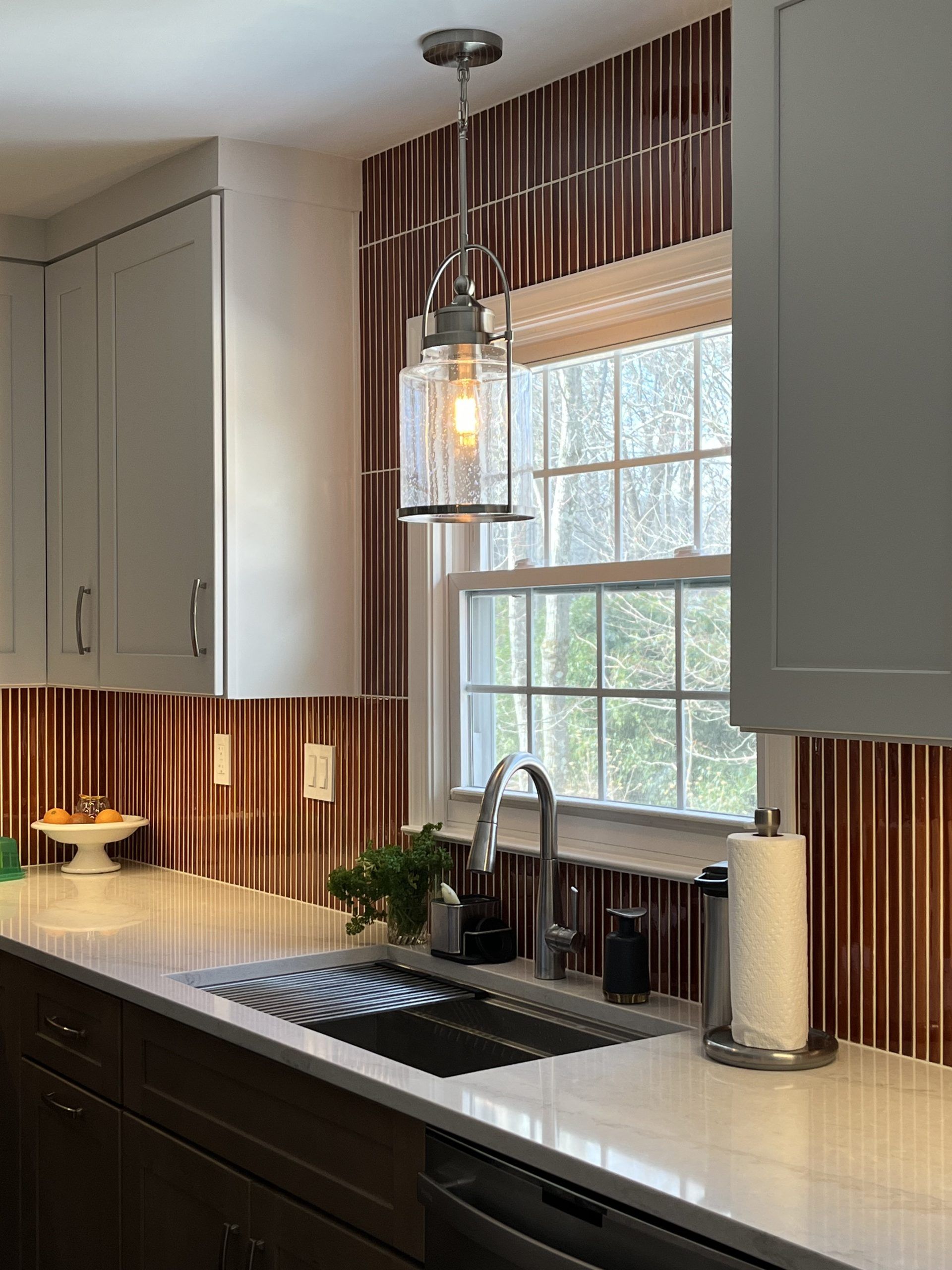 Kitchen with a stainless steel sink, white countertops, and brown tile backsplash. A window is in the background.