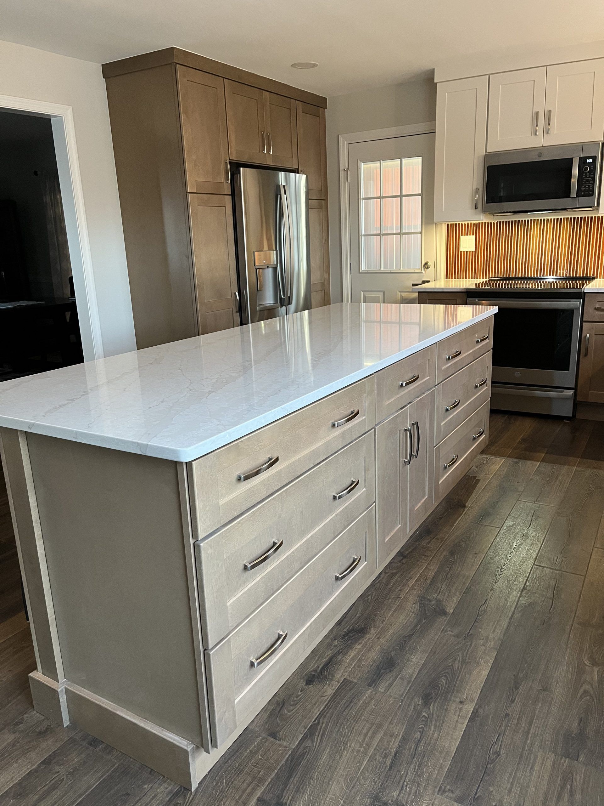 Light-colored kitchen island with drawers and cabinets, light countertop, dark wood floor, cabinets and stainless appliances in background.