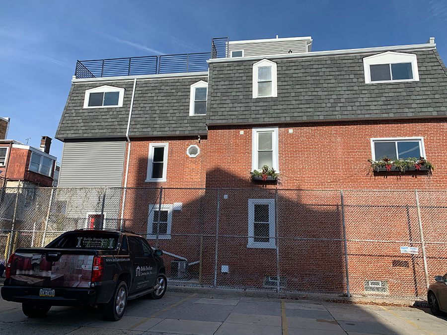 Red brick building with gray roof and windows, a black truck is parked in front of a chain-link fence.