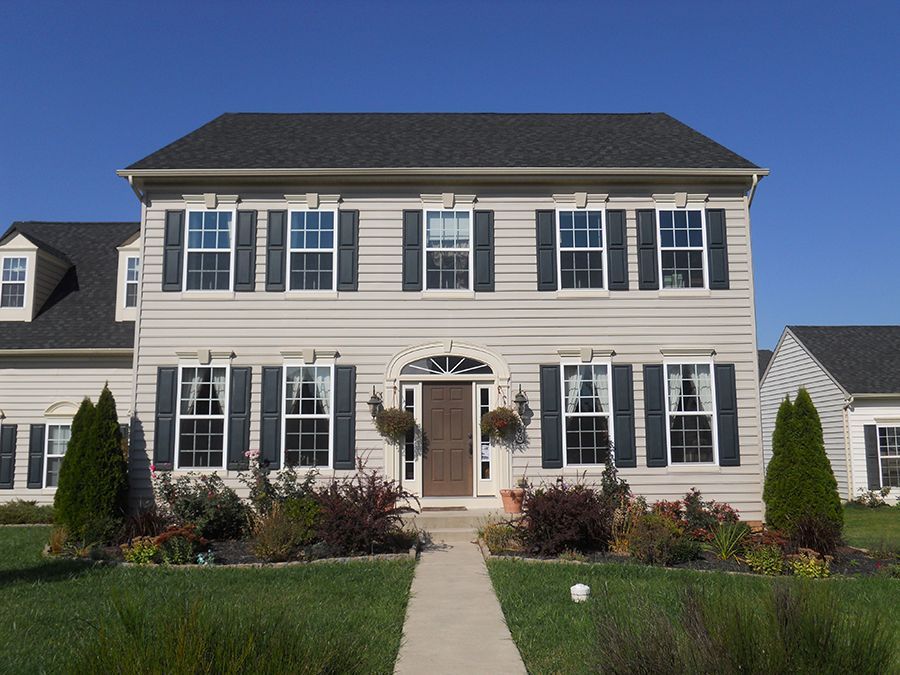 Two-story beige house with black shutters, black roof, and manicured lawn on a sunny day.