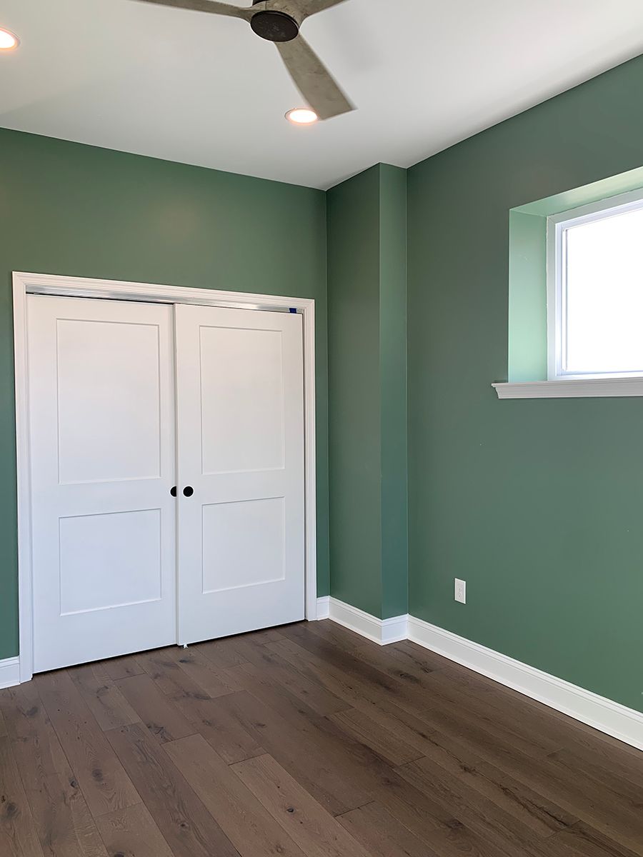 Green bedroom with white closet doors, window, and trim, wood floor, and ceiling fan.